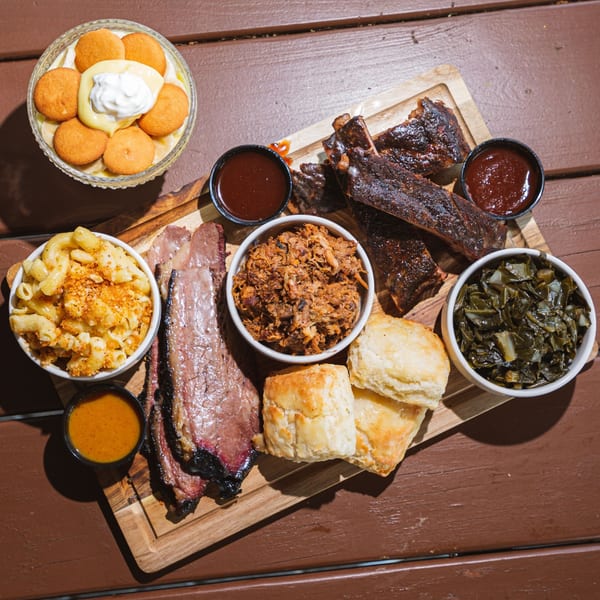 A wooden board filled with Fryer's Roadside BBQ ribs, brisket, pulled pork, mac and cheese, collard greens, biscuits, and banana pudding.