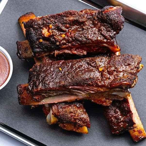 Four large, dark, glazed ribs with a side of barbecue sauce on a black tray.