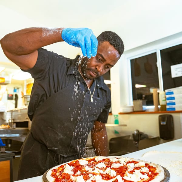 A chef in blue gloves sprinkles seasoning onto a pizza with red sauce and white cheese.