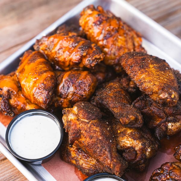 A metal tray filled with Fryer's Roadside fried chicken wings, some sauced, with two dipping cups.